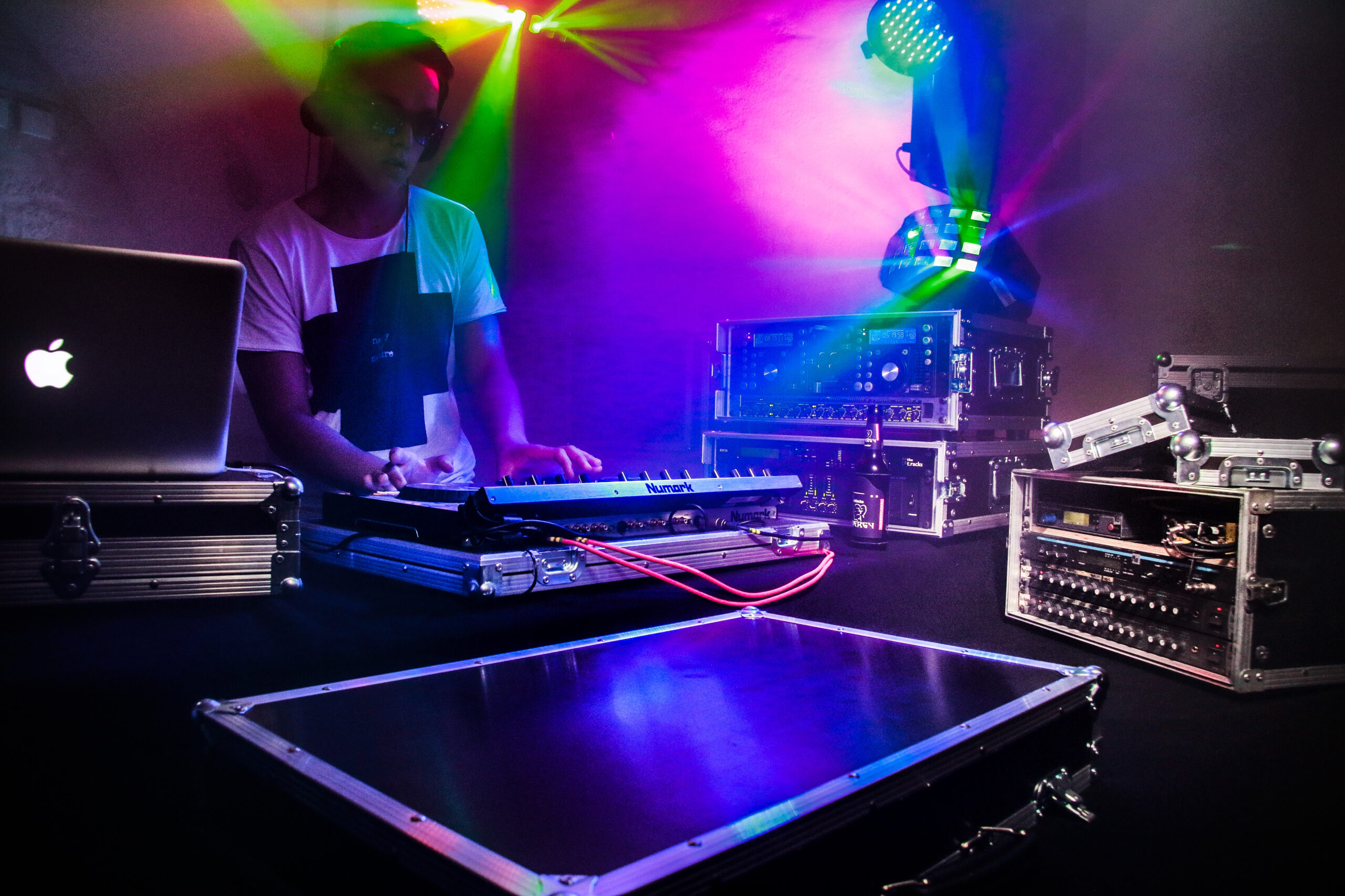 DJ-Performance mit bunten Lichteffekten auf der Bühne. Ein DJ spielt auf einem Keyboard, das auf einem Flightcase steht. Umgeben von professionellem Audio-Equipment, darunter Verstärker und Mischpulte in robusten Transportkoffern.