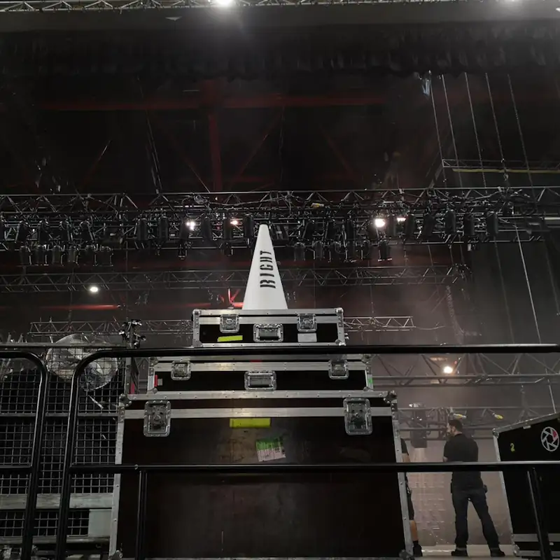 Backstage-/Bühnenszene mit mehreren übereinander gestapelten Flightcases, darauf ein weißer Warnkegel; im Hintergrund Traversensystem mit Lichttechnik, rechts eine Person bei Aufbauarbeiten.