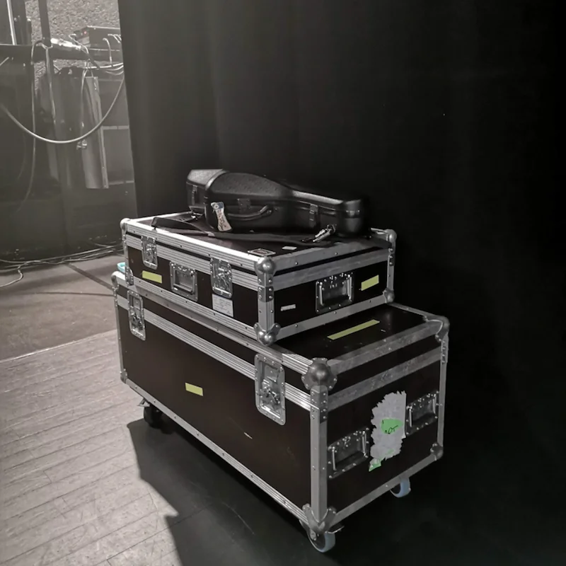 Backstage-Szene am Bühnenrand: zwei verschließbare Flightcases auf Rollen, darauf ein Instrumentenkoffer; links Kabel und Technik, rechts schwarzer Vorhang.