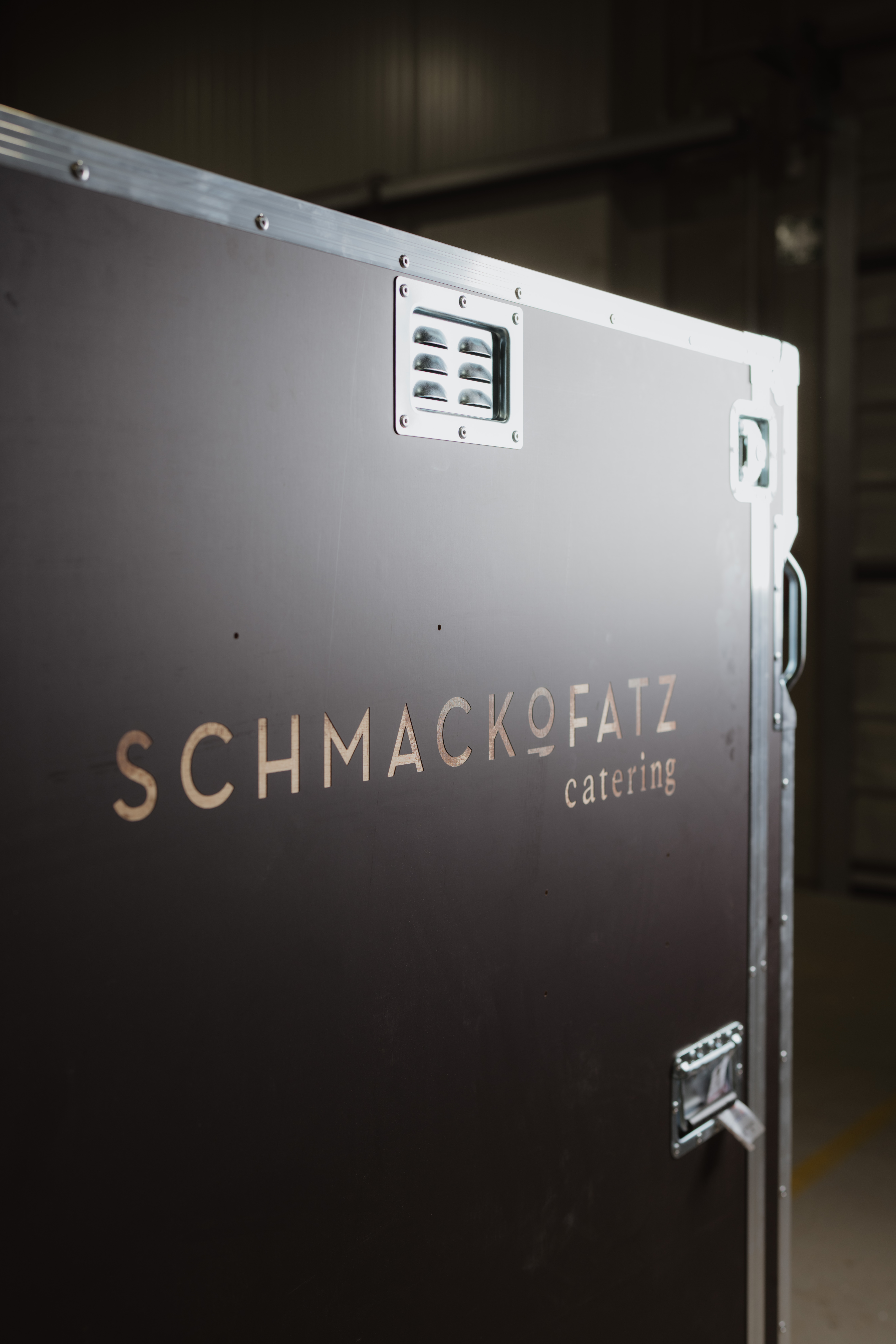 Nahaufnahme eines schwarzen Flightcase mit seitlichem Logo ‚Schmackofatz Catering‘ in einer Halle.